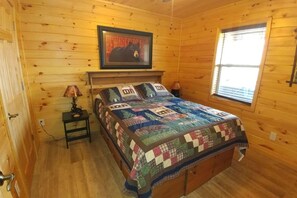 Un dormitorio de cabaña de madera con una cama, una mesita de noche, una lámpara, un cuadro en la pared y una ventana.