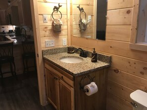 Un baño al estilo de cabaña de madera con lavamanos de encimera de granito, armarios de madera y un espejo sobre el lavamanos.