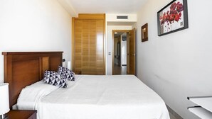 Hotellihuone, jossa on puinen sängynpääty, valkoisella päiväpeitteellä ja kuviollisilla tyynyillä varustettu sänky, yöpöytä lampun kanssa ja seinällä oleva kehystetty, kukkateemainen taideteos.