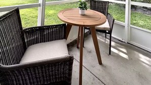 Un porche cubierto con mobiliario de mimbre y una pequeña mesa, con vistas a un área verde y árboles.