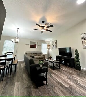 A modern living room with a ceiling fan, a flat-screen TV, a sofa, a coffee table, and a dining area with a table and chairs. - Lakefront Villa in Casselberry (Casselberry)