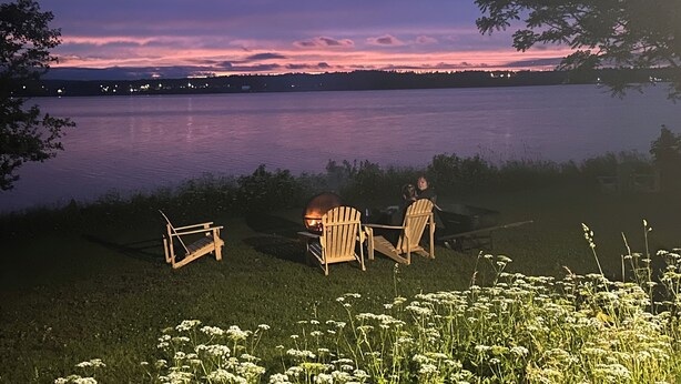 A lakeside scene with two wooden chairs and a small table, a grassy area with flowers, and a sunset over the water. - Rustic waterfront farmhouse on Sydney Harbour, with amazing sunset views. (Point Edward)