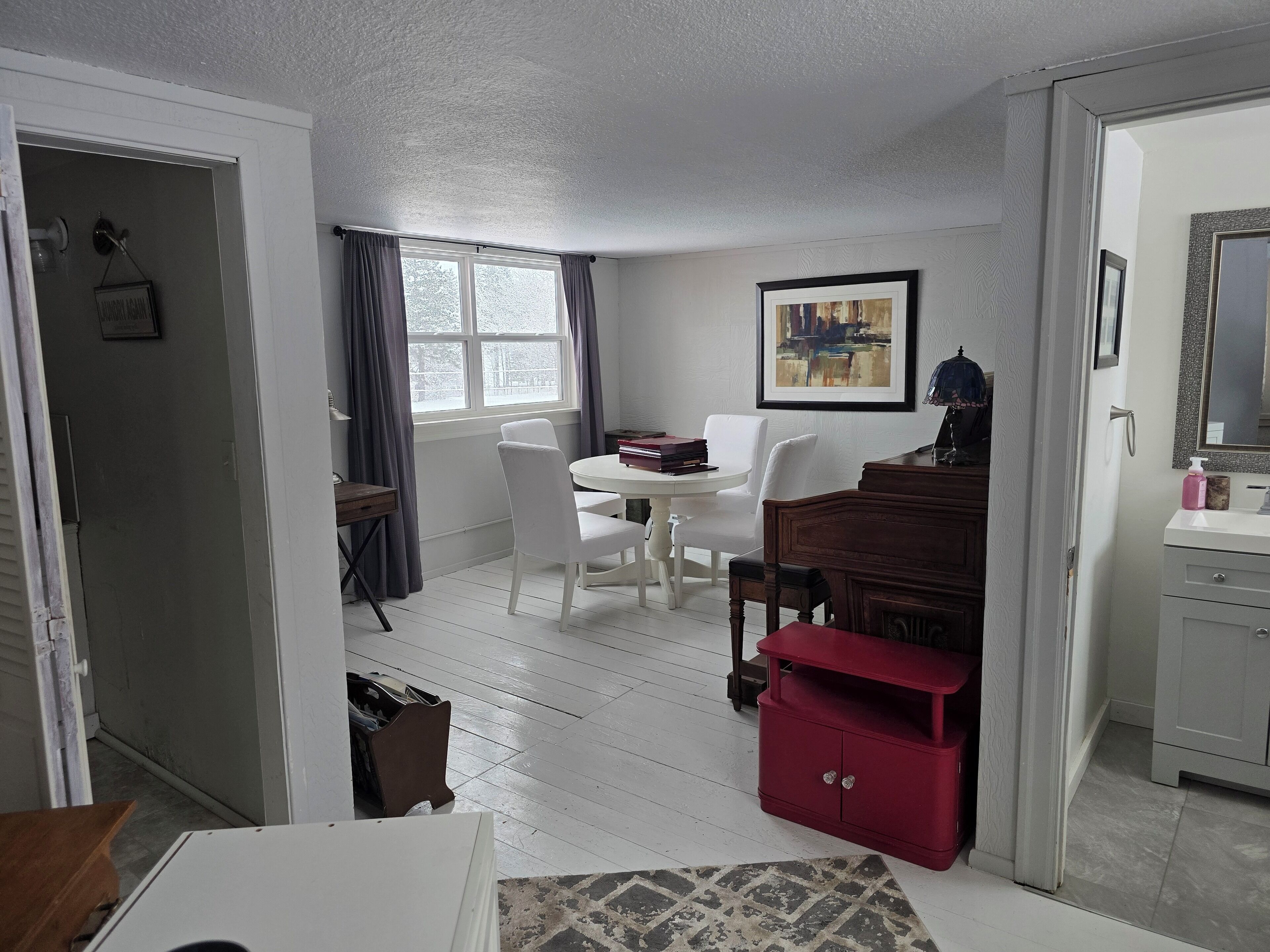 A room with a dining table and chairs, a red cabinet, a white dresser, and a window with curtains.