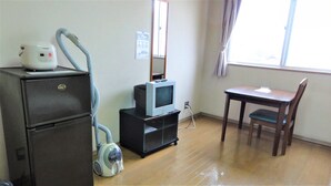 A compact room with a wooden floor, a small desk with a chair, a television on a stand, a vacuum cleaner, and a refrigerator. - Hotel Stay Inn San Annex (Yamagata)