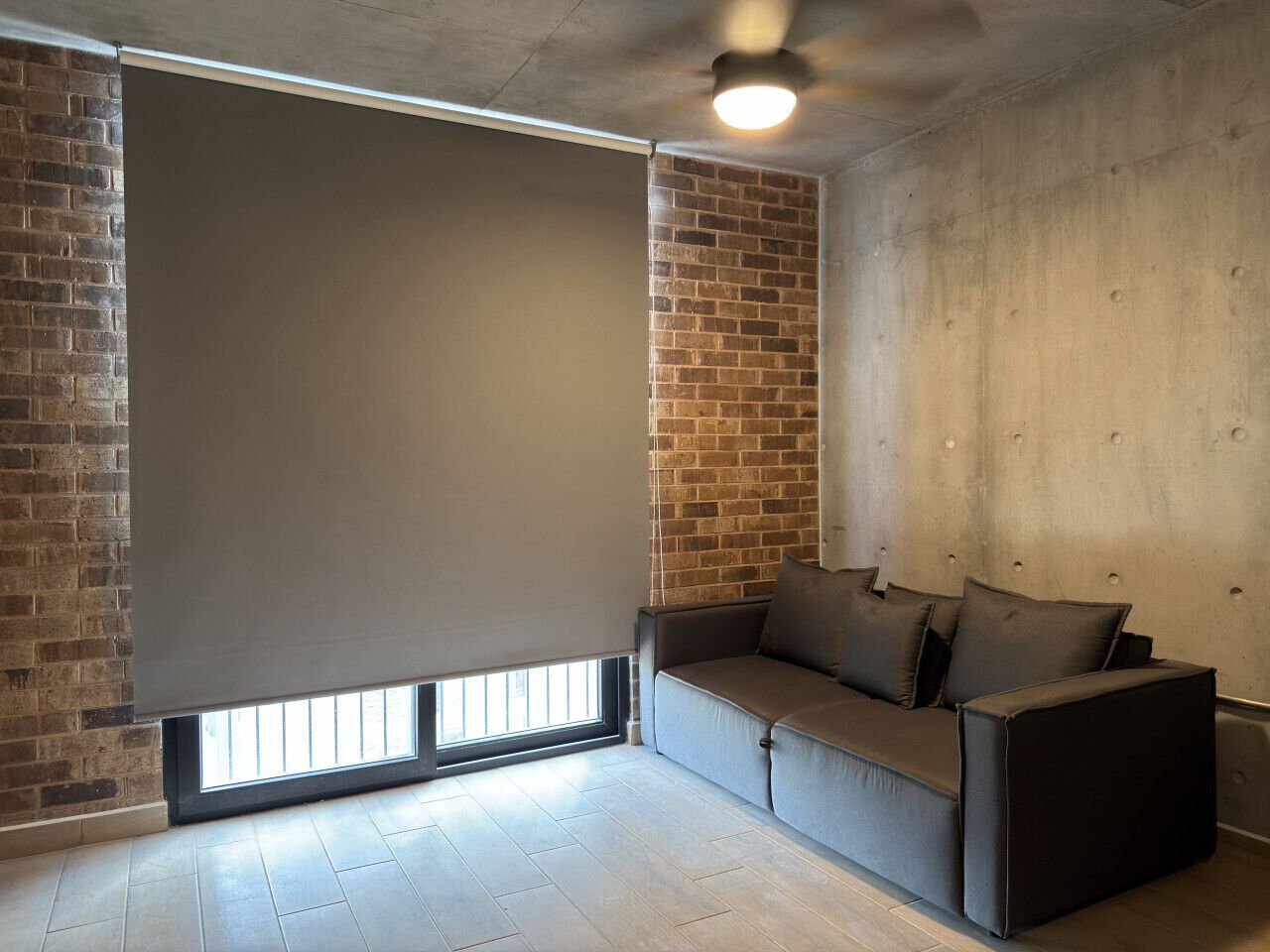 Ein Zimmer mit Backsteinwand, großem Fenster mit Jalousie, einer Couch und einem Deckenventilator.