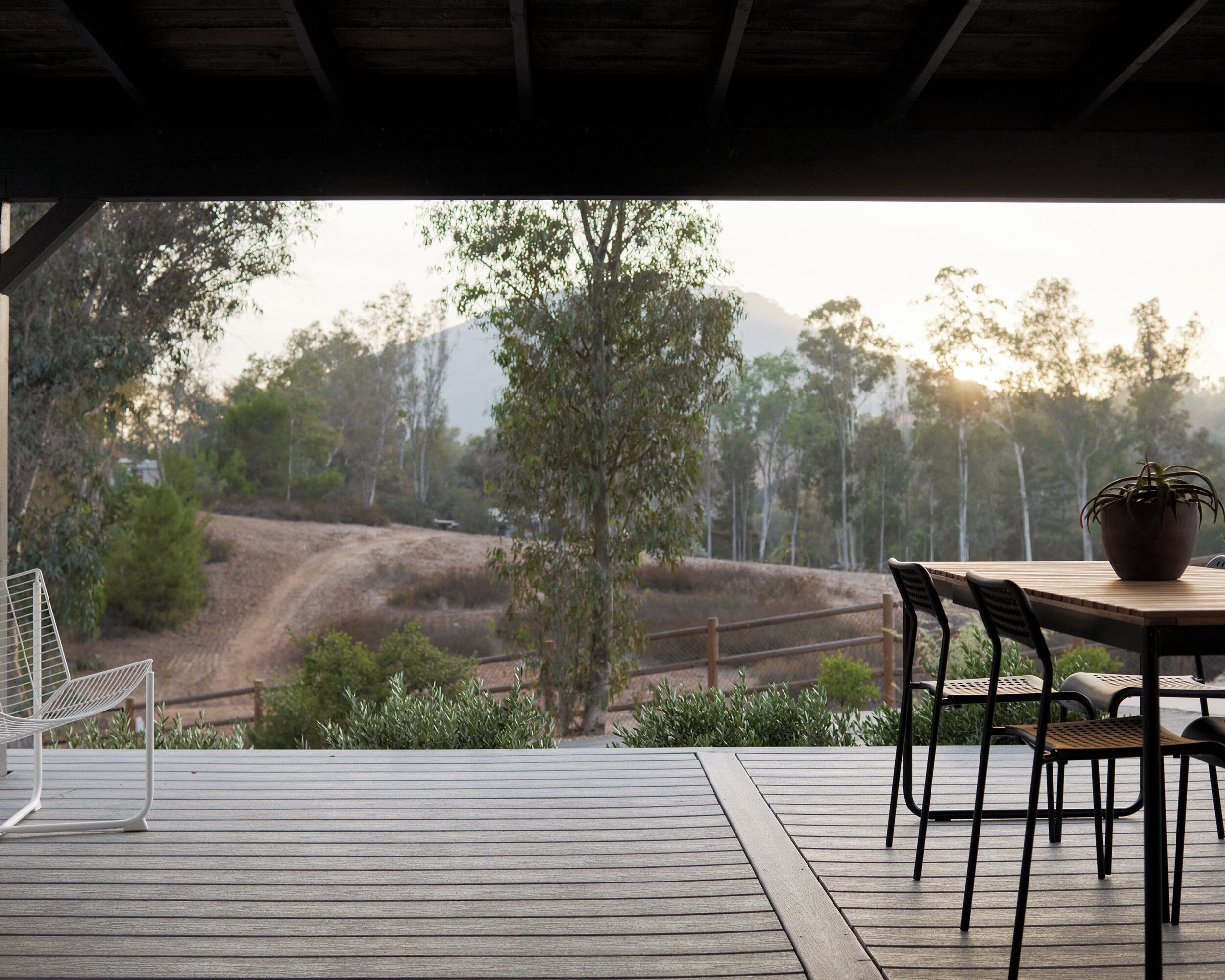 Un terrazzo in legno con tavolo e sedie, affacciato su un sentiero sterrato e alberi.