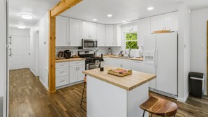 A modern kitchen with white cabinets, a wooden beam ceiling, a central island with a wine glass, and a window with a view of greenery. - Stylish Knoxville Retreat w/ Media Room & Patio (Knoxville)