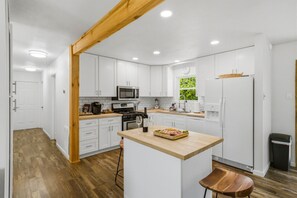 A modern kitchen with white cabinets, a wooden beam ceiling, a central island with a wine glass, and a window with a view of greenery. - Stylish Knoxville Retreat w/ Media Room & Patio (Knoxville)