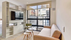 Een moderne woonkamer met een flatscreen televisie, een bank, een eettafel met stoelen en een groot raam met uitzicht op gebouwen.