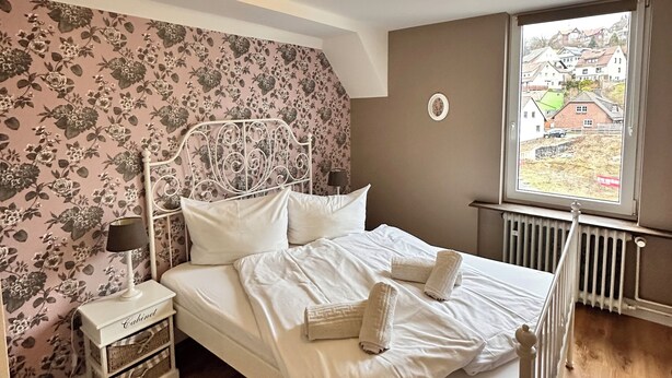 Quarto com cama, mesa de cabeceira, abajur, cômoda e janela com vista para casas.