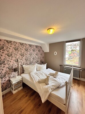 A bedroom with a bed, a nightstand, a lamp, a dresser, and a window with a view of houses. - Our fairytale house in Braunlage vacation apartment Alice in Wonderland (Braunlage)