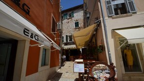Una stretta via con un ristorante chiamato 'Marinero' sullo sfondo, una insegna per 'E&G planet' a sinistra e una scultura in legno raffigurante una donna che tiene una lanterna in primo piano.