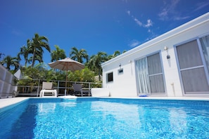 A swimming pool with clear blue water, surrounded by a white building with large windows and a covered area with chairs and an umbrella. - Crystal Villa Imgya <miyakojima> (Miyakojima)
