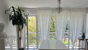 Tilava ruokailutila, jossa on pitkä pöytä, valkoiset verhot, kattokruunu ja ruukussa oleva kasvi.