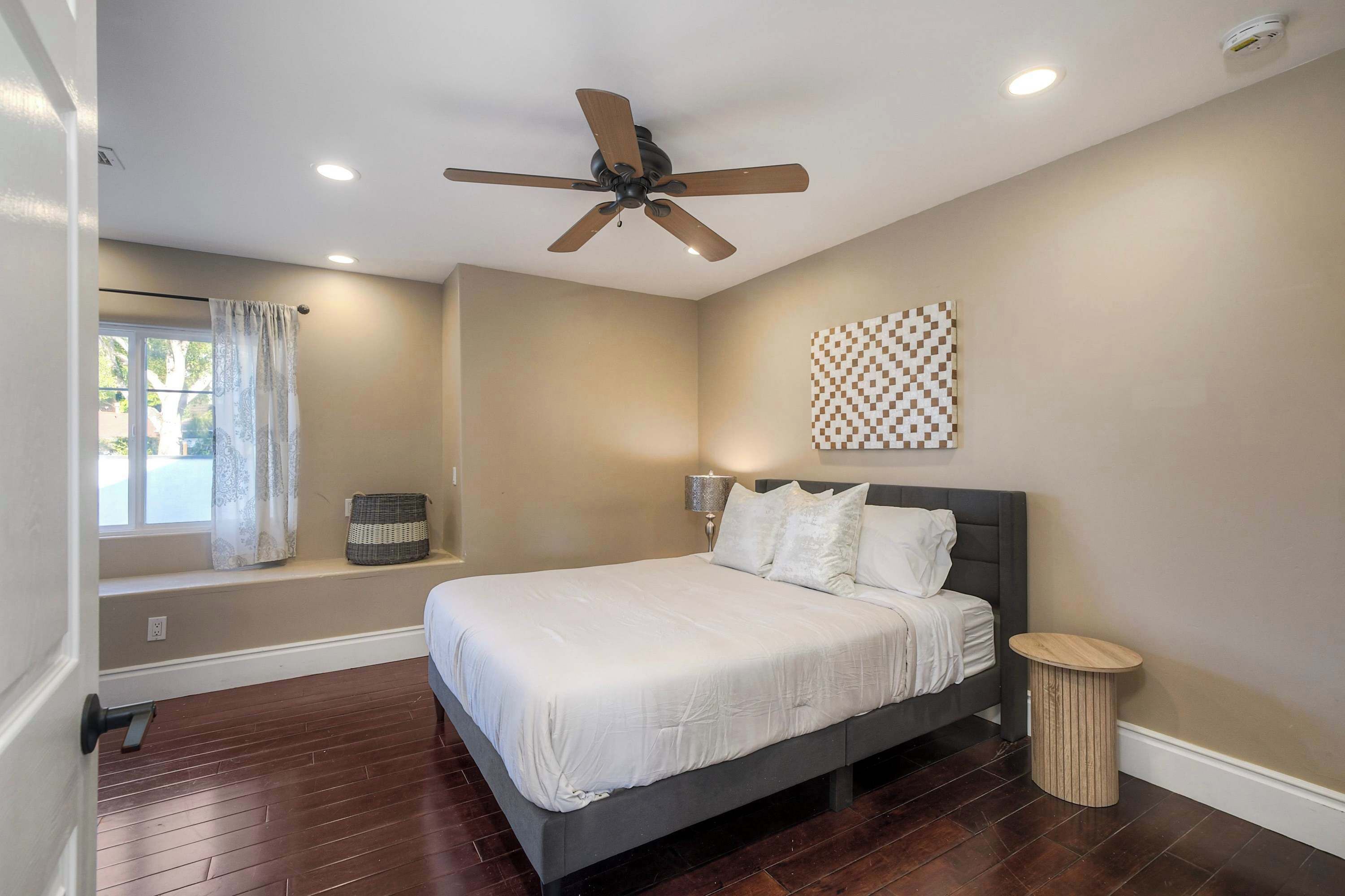 A bedroom with a bed, a ceiling fan, a window with curtains, a wall-mounted lamp, and a small round table.