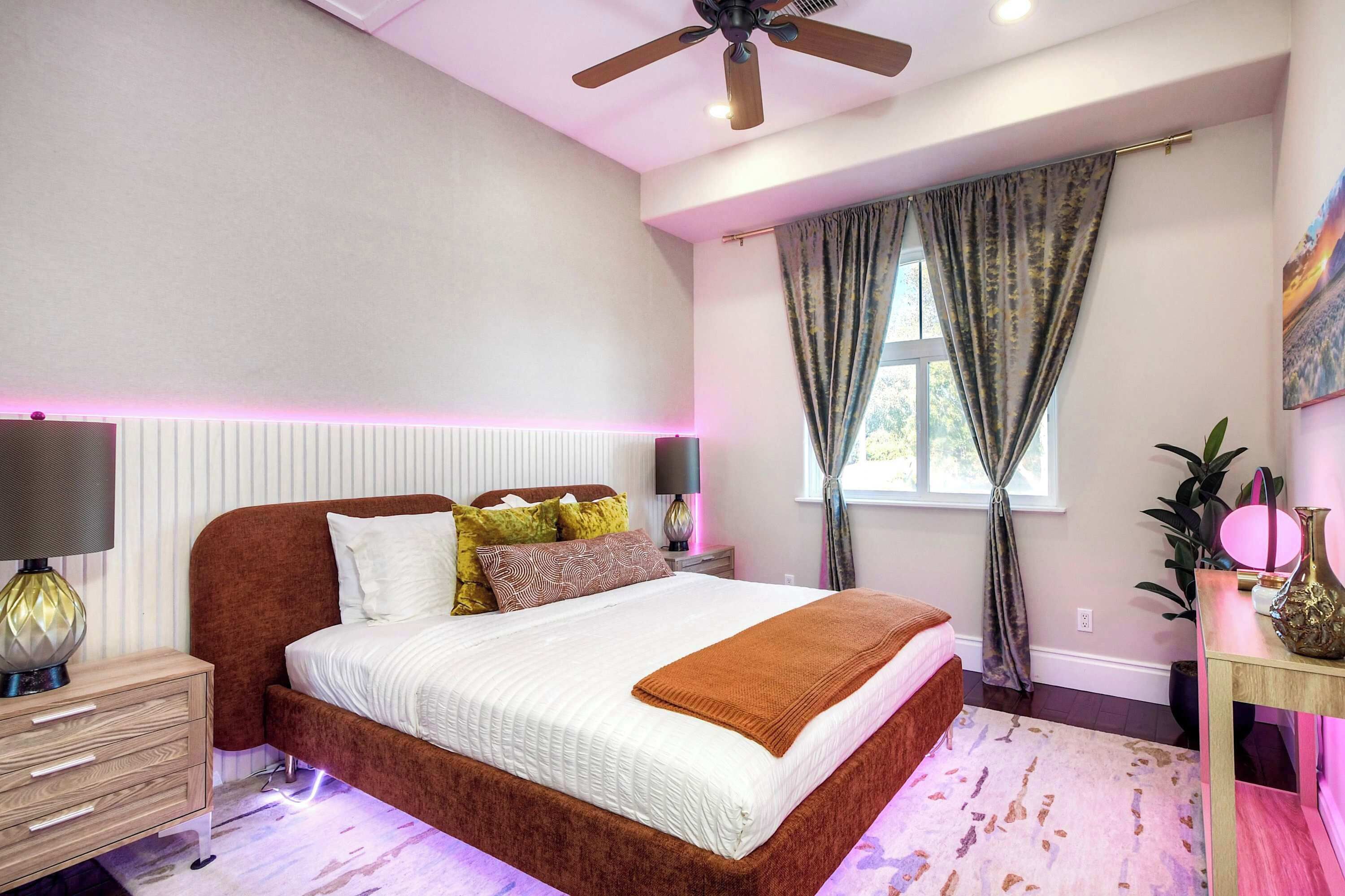 Une chambre à coucher comprenant un lit, des tables de chevet, un ventilateur de plafond, une fenêtre avec des rideaux et une petite table de nuit avec une lampe.