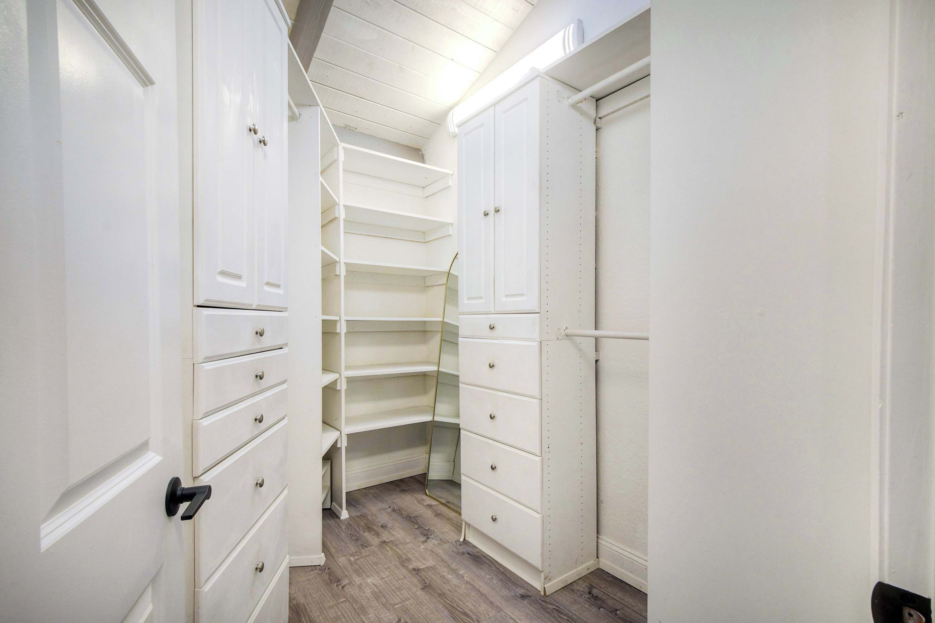 A narrow hallway with white storage cabinets and drawers, wooden flooring, and a white ceiling with exposed beams.