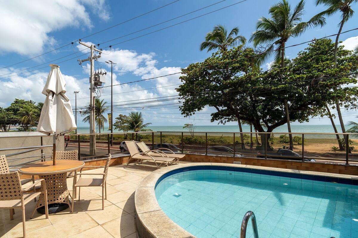 Área da piscina com espreguiçadeiras e uma mesa, com vista para uma praia com palmeiras e céu claro.