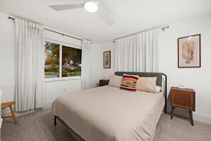 A bedroom with a bed, a bedside table, a nightstand, a ceiling fan, a window with curtains, and a framed picture on the wall. - Sun-Kissed Modern Oasis with Private Heated Pool (Fort Myers)