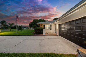A residential garage with a driveway leading to a house, an American flag, and a sunset sky. - Sun-Kissed Modern Oasis with Private Heated Pool (Fort Myers)