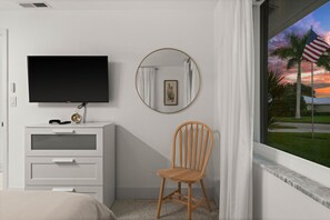 A bedroom with a bed, a TV, a dresser, a chair, a round mirror, and a window with a view of palm trees and a sunset. - Sun-Kissed Modern Oasis with Private Heated Pool (Fort Myers)