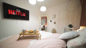 Ein Schlafzimmer mit einem Bett, einem Nachttisch, einem an der Wand montierten Fernseher, der 'NETFLIX' zeigt, und einem Fenster mit Vorhängen.