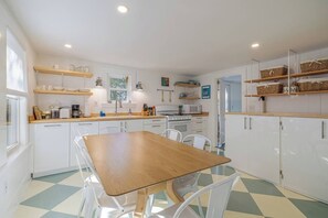 A kitchen with white cabinets, a wooden table, and white chairs. There are shelves with kitchenware and a window above the sink.