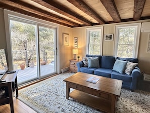 A cozy living room with a blue sofa, wooden coffee table, and a large window with a view of trees. - 3BR Cottage in Center of Acadia! [Country Cottage] (Mount Desert)