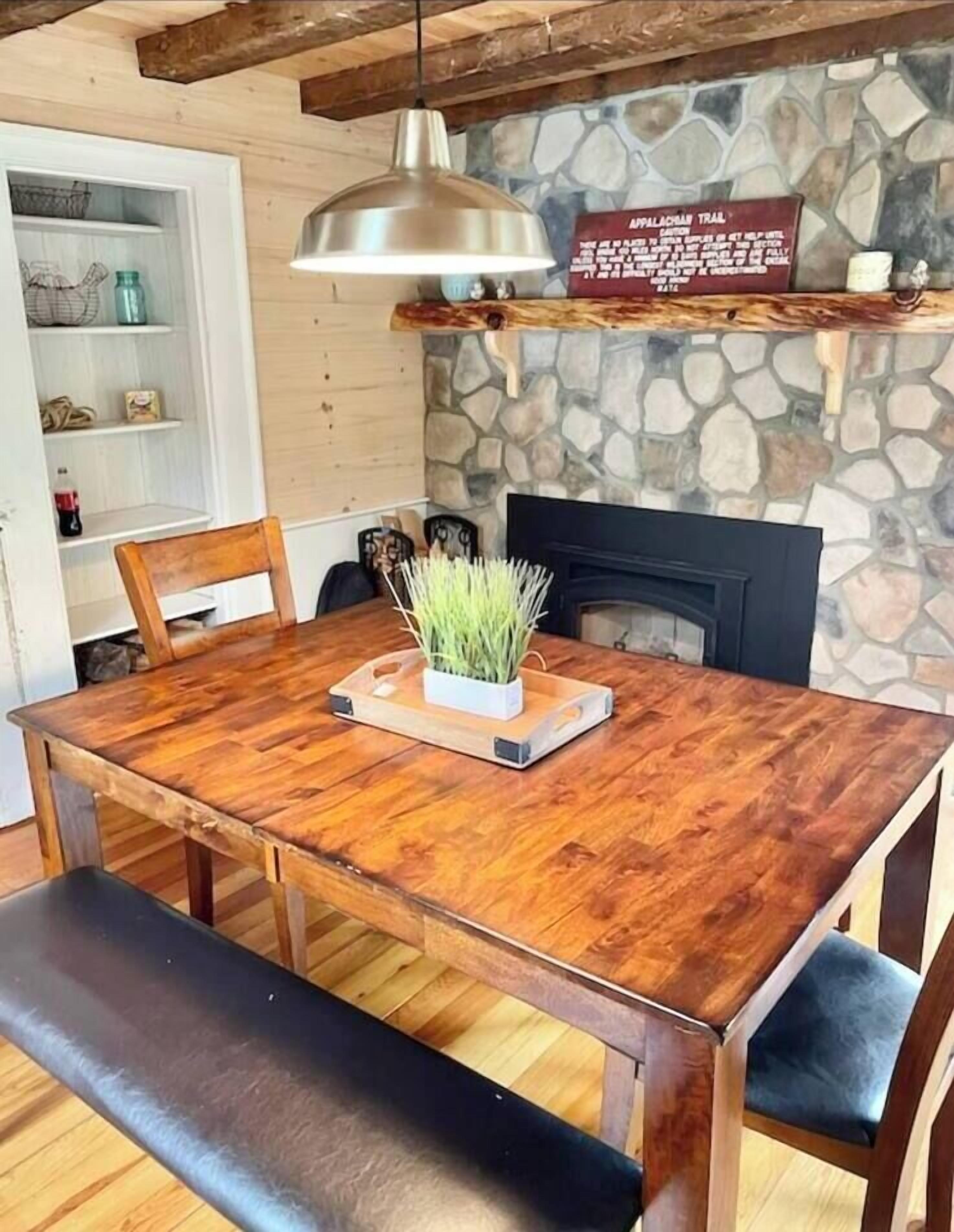 Een houten eettafel met stoelen, een hangende hanglamp, een stenen muur en een open haard.