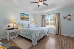 A bedroom with a bed, two bedside tables with lamps, a ceiling fan, a window with curtains, and a wall-mounted painting. - Pup friendly, spacious 4BR home, gourmet kitchen, golf cart avail in Waveland. (Waveland)