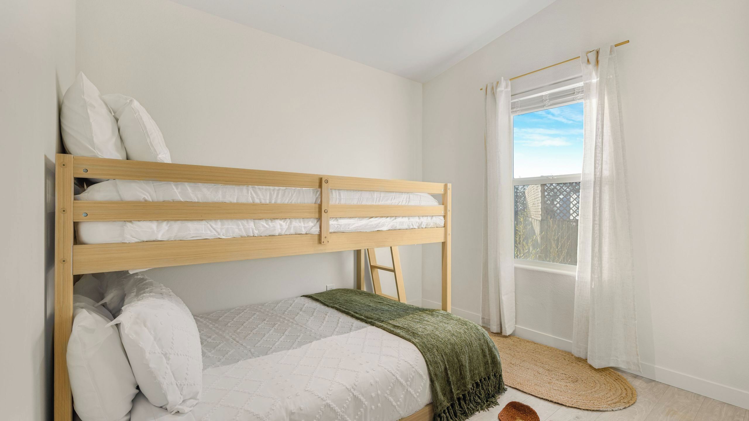 A bunk bed with white bedding, a green throw blanket, a window with white curtains, and a wooden floor.
