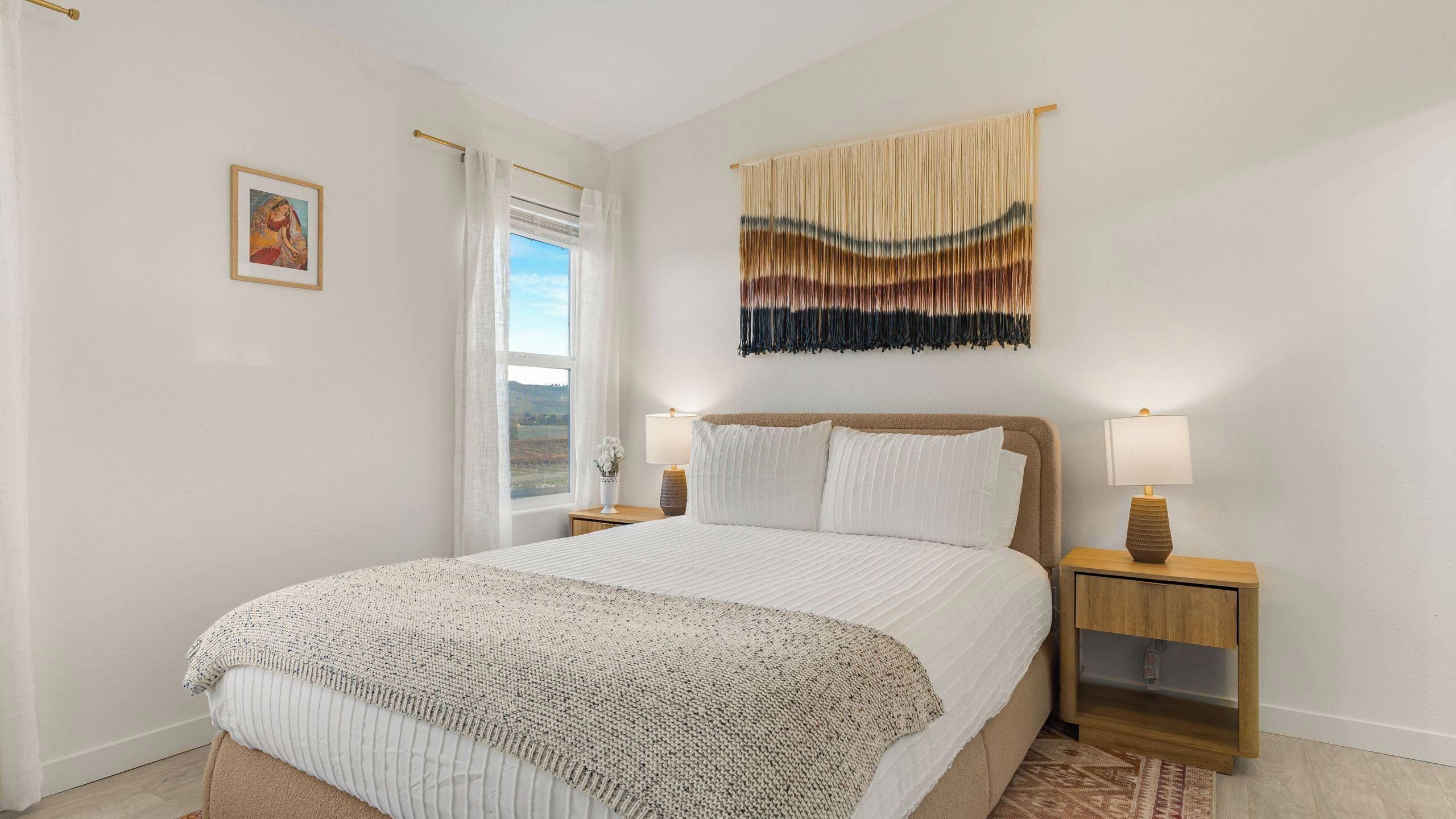Un dormitorio ordenado con una cama grande, dos mesitas de noche con lámparas, un ventanal con cortinas y un adorno en la pared.