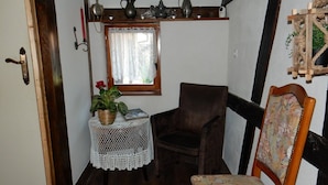 Une petite pièce avec une chaise en bois, une table recouverte d’une nappe en dentelle, une plante en pot et une fenêtre avec un rideau.