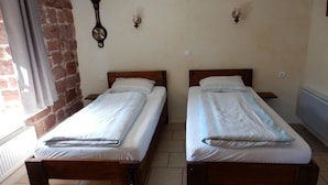 Deux lits en bois, recouverts de draps blancs, dans une pièce dont un mur est en briques et où l’on aperçoit une horloge.