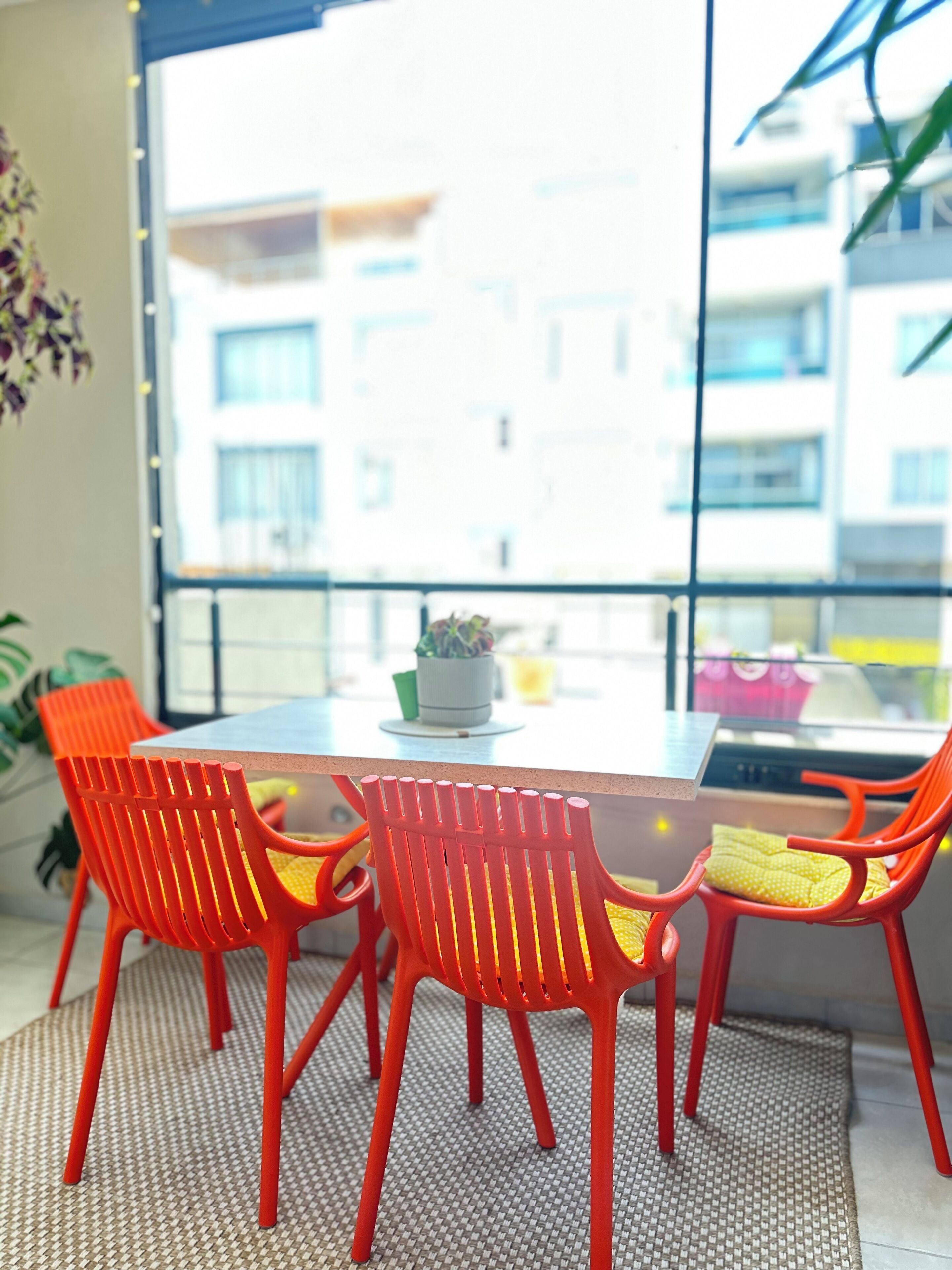 Een eetruimte met een tafel en vier oranje stoelen, een potplant en uitzicht op gebouwen door een groot raam.