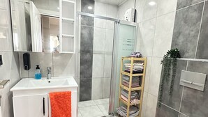 Et moderne badeværelse med en hvid vask, spejl og toilet. Der er røde måtter på gulvet, en brusekabine i glas og en træhylde med håndklæder.