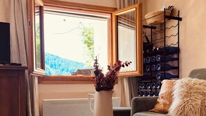 A cozy living room with a wooden table, a comfortable sofa, a small stool, and a window with curtains. - Grange pour 6 personnes avec studio indépendant (Beuil)
