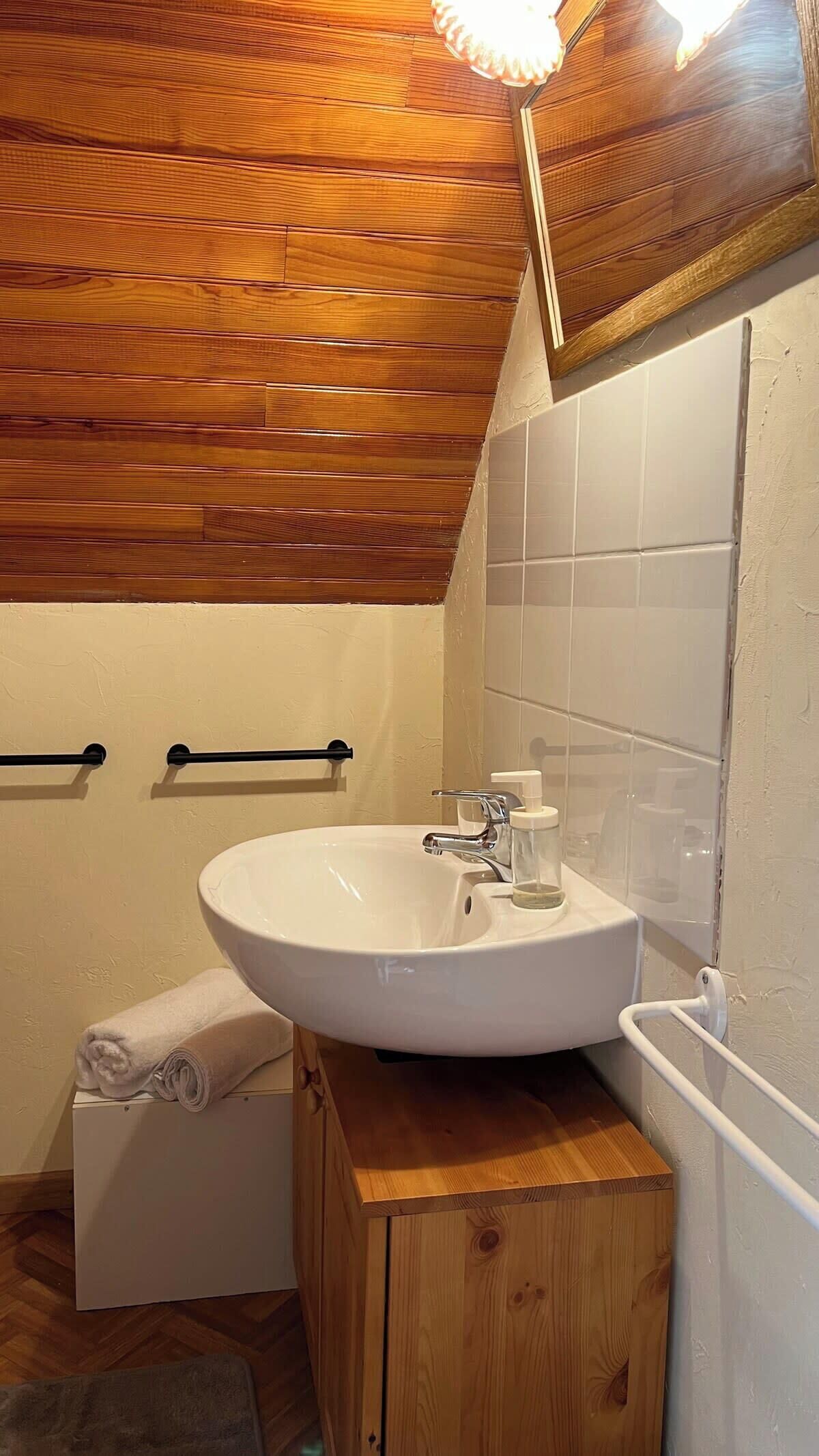 A bathroom with a wooden ceiling, white tiles, a sink with a soap dispenser, and a towel on a wooden cabinet.