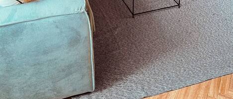 Una sala de estar con piso de madera en espiga, un sofá azul, una mesa de centro de vidrio y un cuadro en la pared.