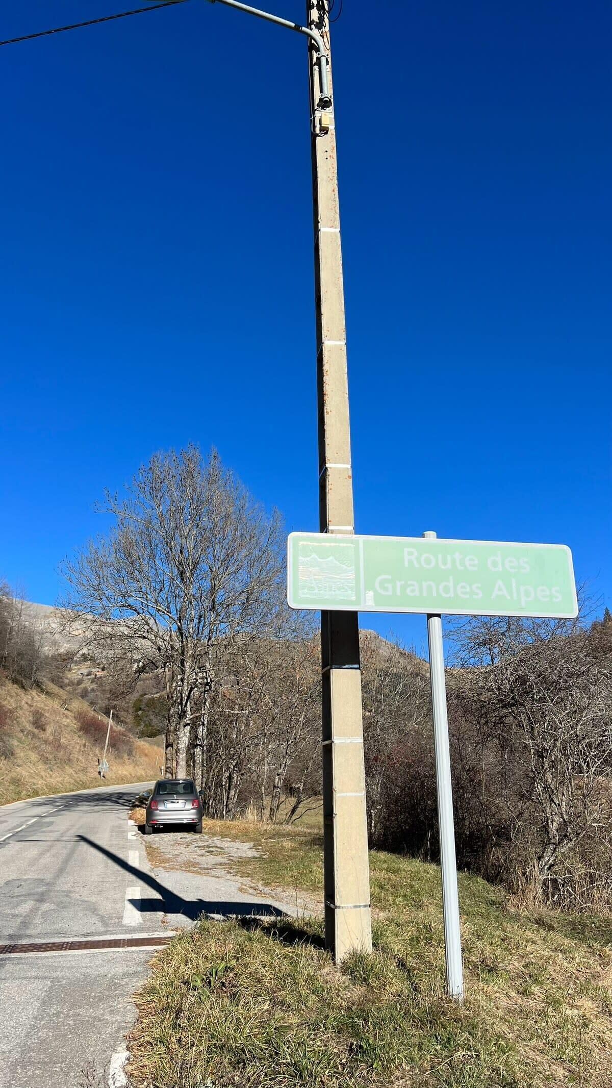 Segnaletica stradale per la Route des Grandes Alpes, con un cielo azzurro limpido e un'auto parcheggiata sul lato della strada.