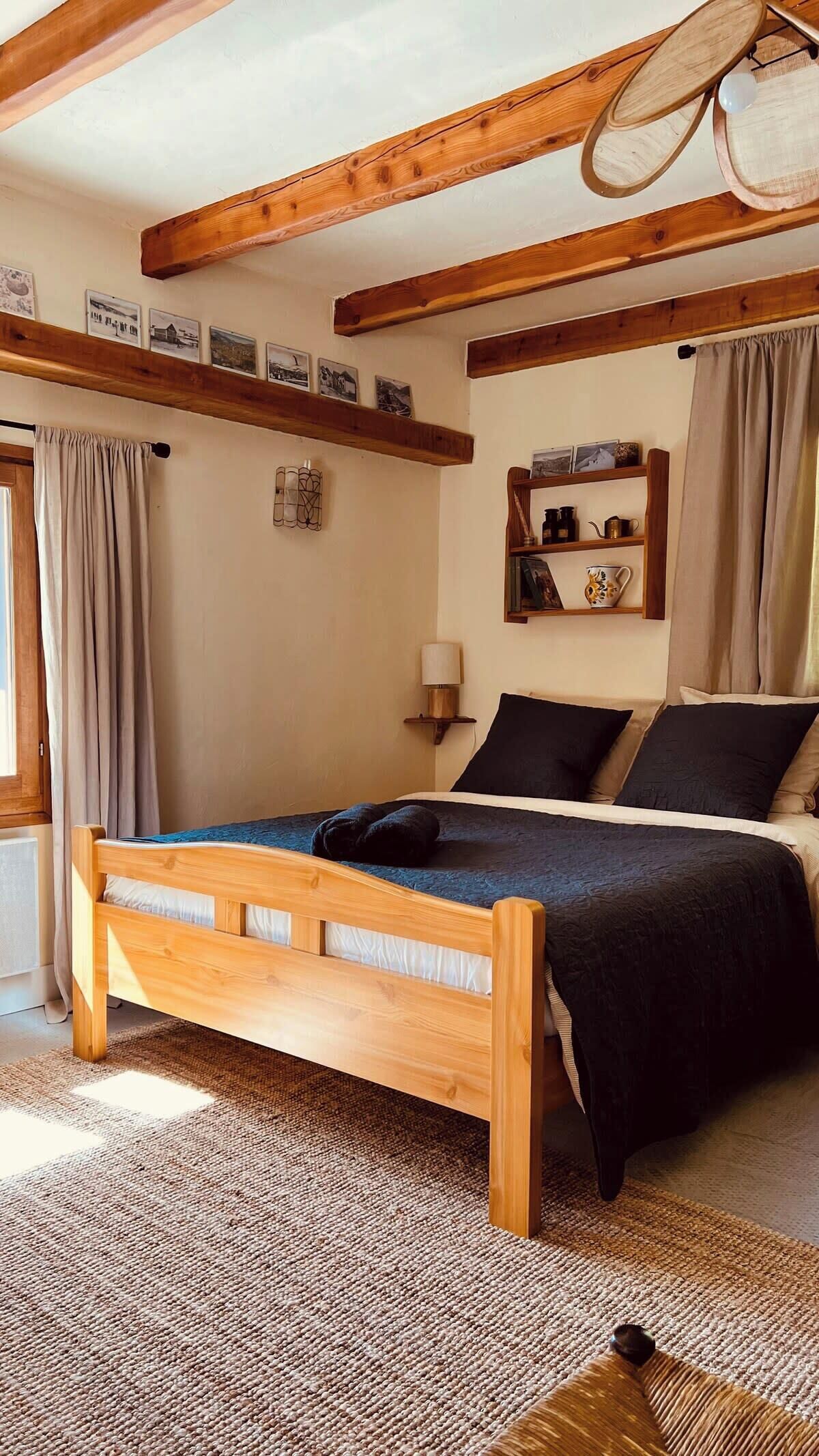 Un lit en bois recouvert d’une couverture bleue, dans une pièce dont les poutres apparentes sont en bois. On y trouve également une étagère avec des photos et une lampe, ainsi qu’une fenêtre avec des rideaux.