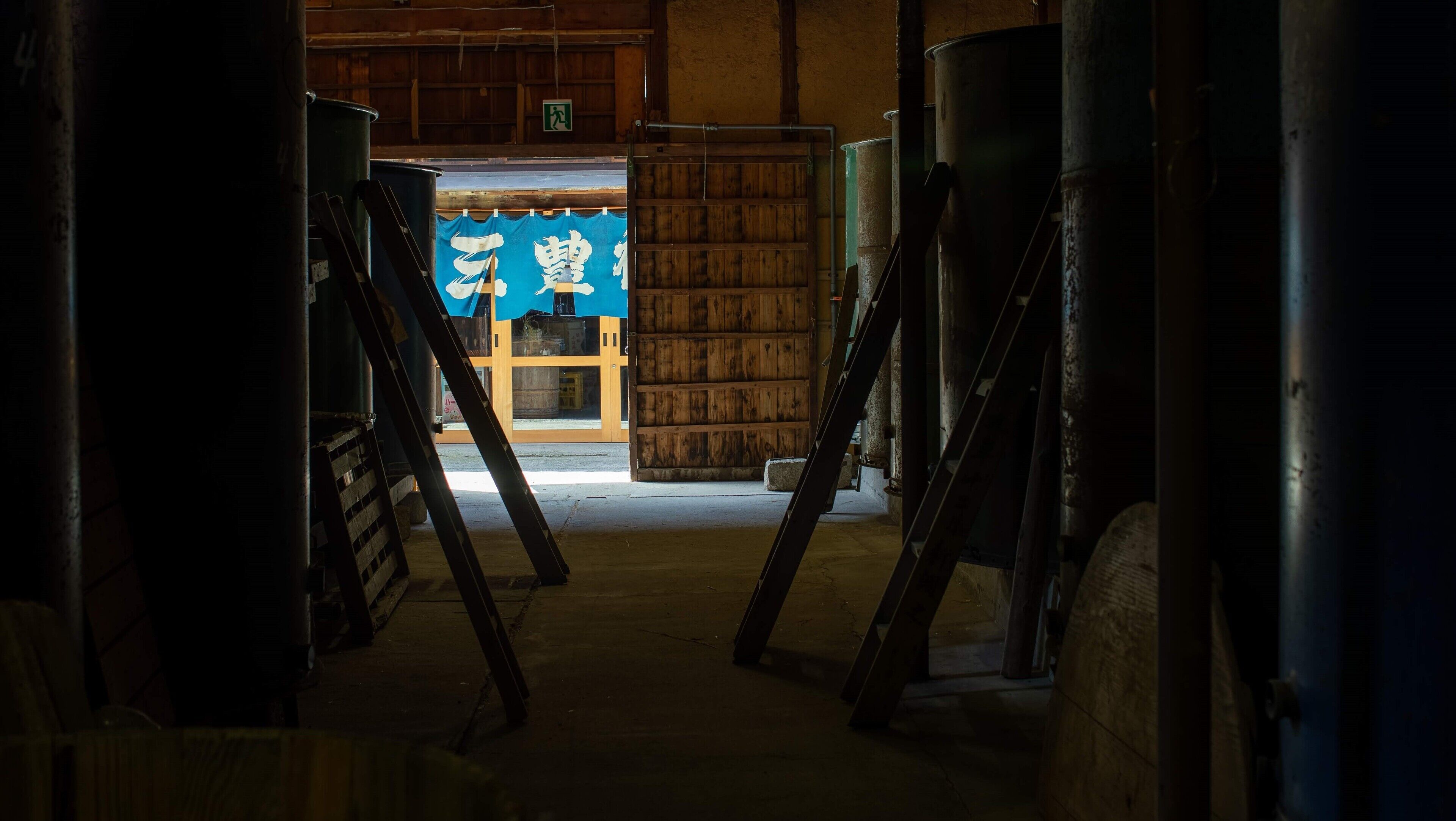A corridor with wooden doors and metal structures, leading to a room with a blue sign.