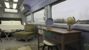 A train compartment with a bed, a desk with a globe and mirror, a chair, and a window with a view of the outside.