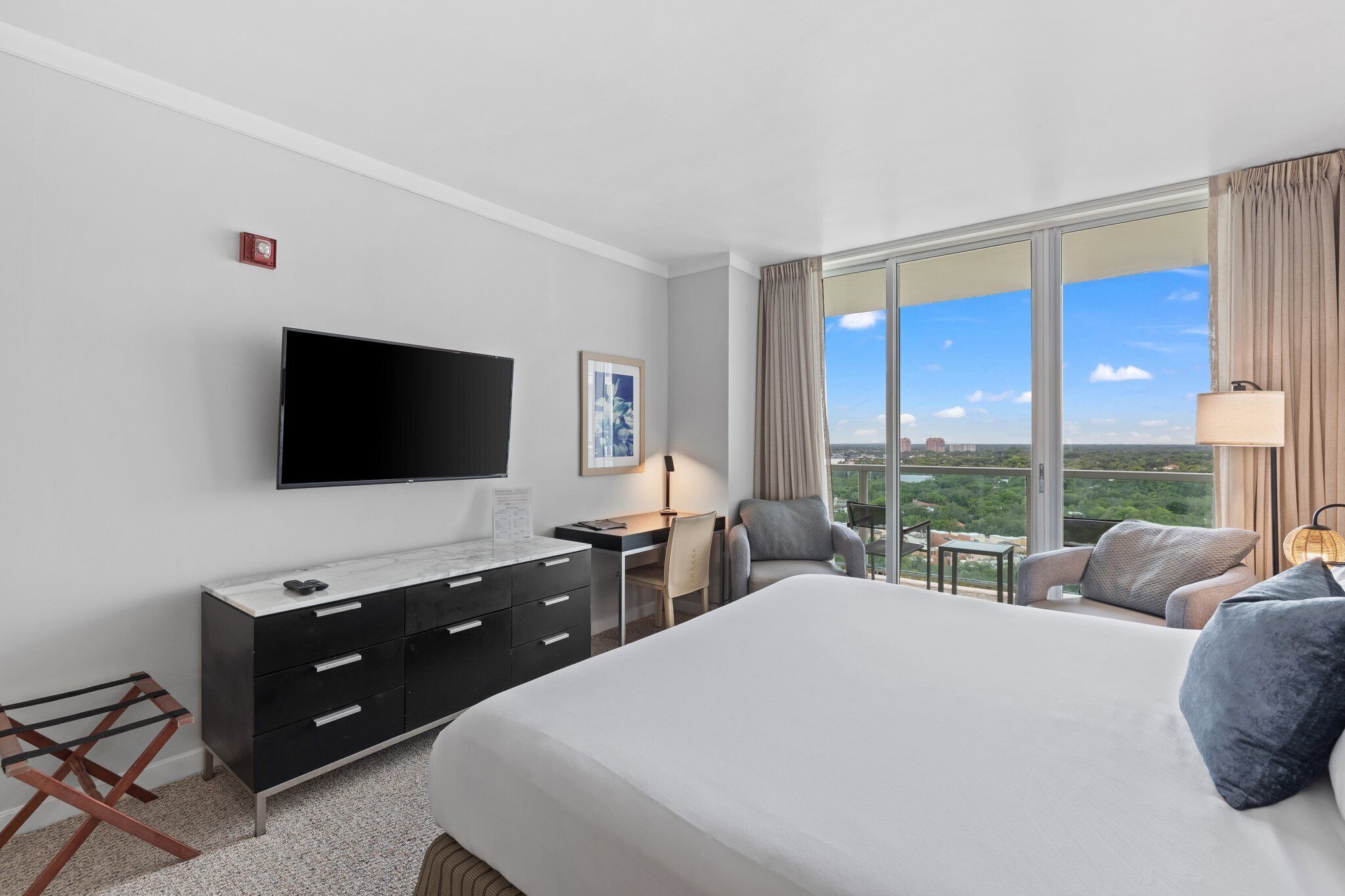 A hotel room with a large bed, a desk, a chair, a TV, and a view of the city.