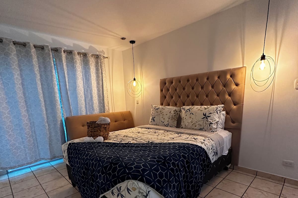 A bedroom with a tufted headboard, a patterned bedspread, and a bedside table with a lamp.