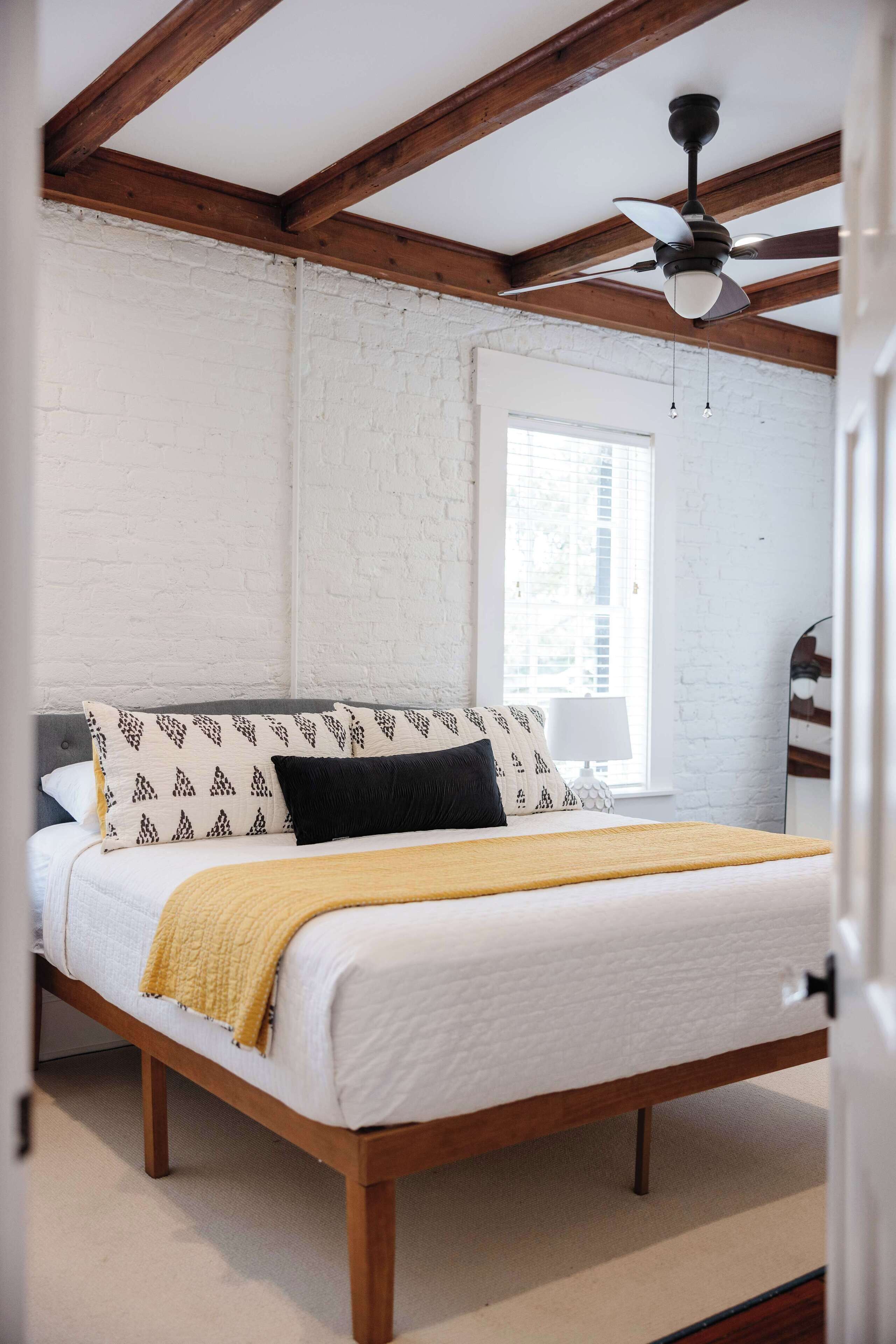 Quarto com cama de madeira, paredes brancas, ventoinha no teto e janela com persianas.