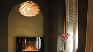 A cozy room with a fireplace, two white cushioned chairs, a woven floor lamp, and a potted plant by the window.