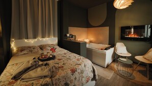 A modern bedroom with a large bed, a sitting area with a chair, a bathtub, and a TV mounted on the wall.