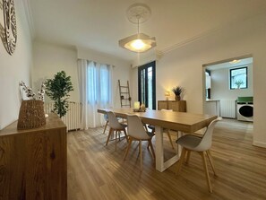 Uma área de refeições moderna com uma mesa e cadeiras de madeira, um lustre pendurado e vista para uma cozinha com uma máquina de lavar.