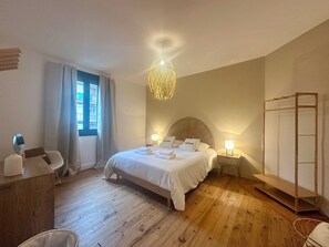 Quarto com piso de madeira, cama com roupa de cama branca, mesa de cabeceira com abajur, janela com cortinas e luminária pendente.